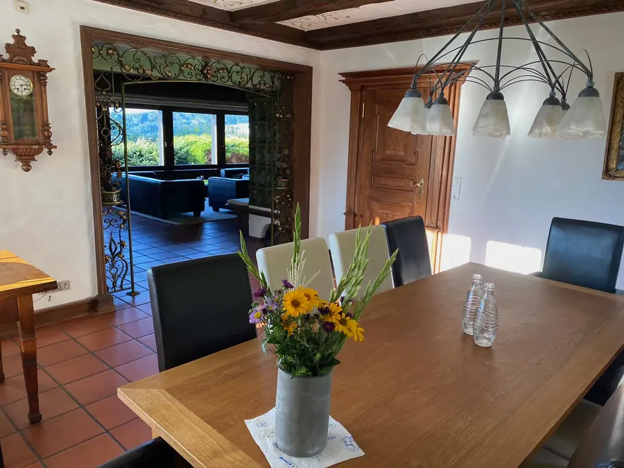Blick vom Esszimmer mit Blumenstrauß durch die geschnitzte Tür ins Wohnzimmer mit Fensterfront zum Tal