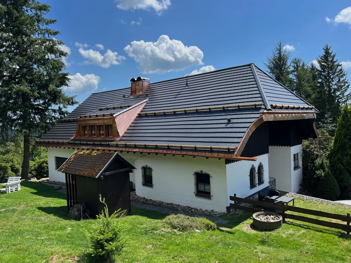 Die Villa im Sommer: weißes Haus mit dunklem Dach und Kupferkanten auf der Wiese, Tannen ringsum, Wolken am blauen Himmel